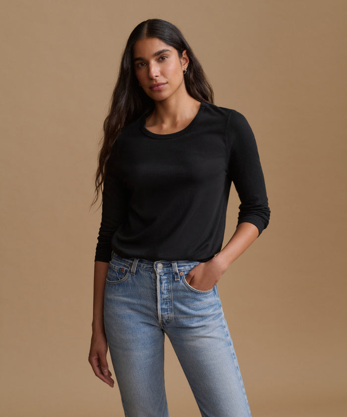 A woman modeling a black long-sleeve crewneck top paired with light-wash blue jeans, standing against a solid warm beige studio background.