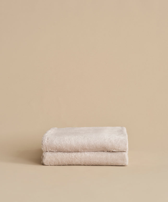 Two neatly folded, super-soft Loom Bath Sheets in beige are stacked atop each other against a plain beige background.