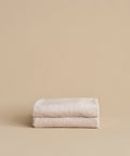 Two neatly folded, super-soft Loom Bath Sheets in beige are stacked atop each other against a plain beige background.