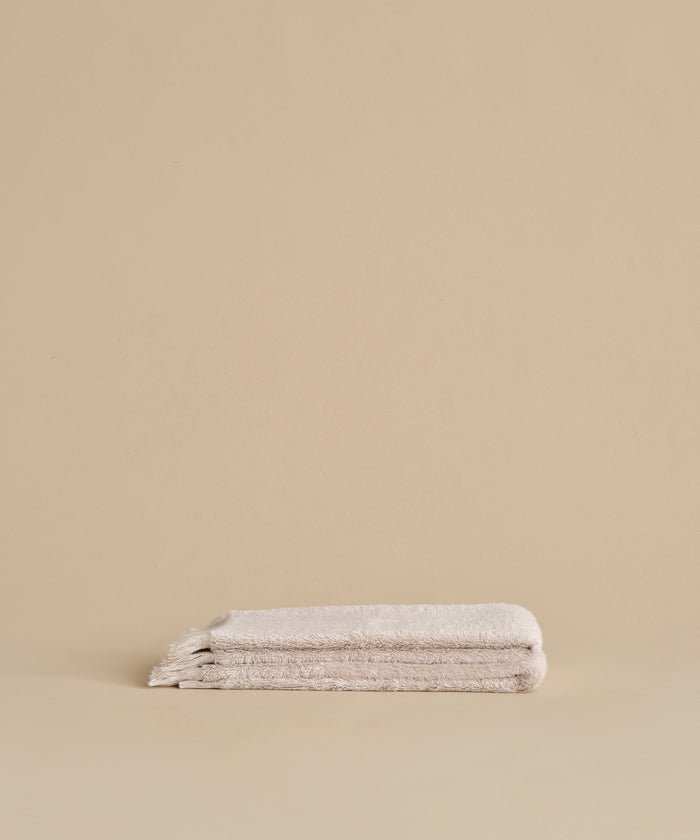 Two neatly folded Loom Hand Towels are stacked on a matching beige surface with a plain beige background, creating a serene bathroom essential display.