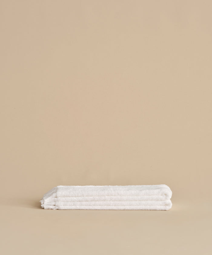 The Loom Hand Towel, ultra-soft and neatly folded in classic white, is displayed on a smooth beige surface with a coordinating beige background—an essential for any bathroom.