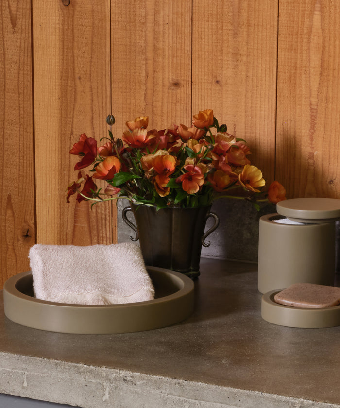 A Loom Hand Towel rests in a round tray beside a metal vase with orange and yellow flowers, a lidded container, and a soap dish—creating an inviting bathroom display on the gray countertop by the wooden wall.
