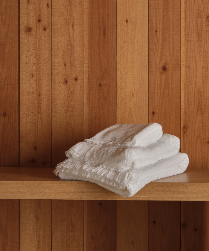 A trio of Loom Hand Towels, super-soft and white, neatly folded on a wooden shelf with vertical wood paneling behind—a true bathroom essential.