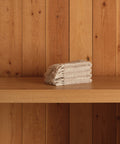 A neatly folded stack of super-soft beige Loom Wash Cloths sits on a wooden shelf with vertical wooden panels in the background—a true bathroom essential.