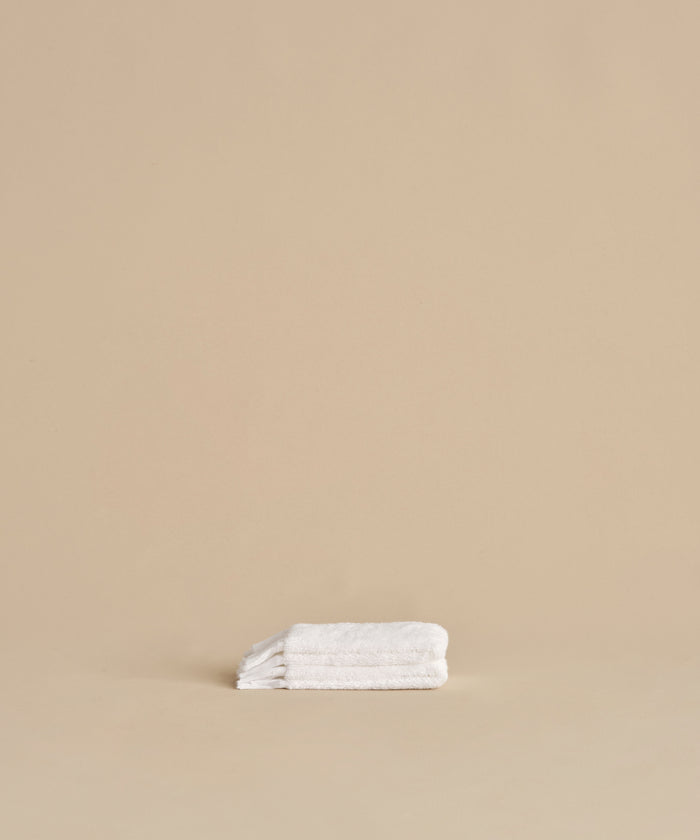 The Loom Wash Cloth, made from white cotton modal, is neatly folded on a beige surface with a plain beige background, showcasing a minimalist and clean bathroom essential.