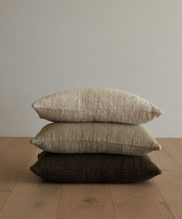 The Luna Lumbar Pillow, handwoven in cream, beige, and dark brown neutral shades, is stacked with two other textured throw pillows on a light wood floor against a plain light-colored wall.