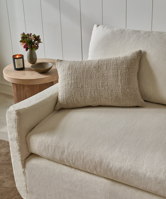 A beige sofa with the Luna Lumbar Pillow in textured tan sits beside a round wood side table displaying a lit candle, a small flower vase, and a decorative bowl, all against a white paneled wall.