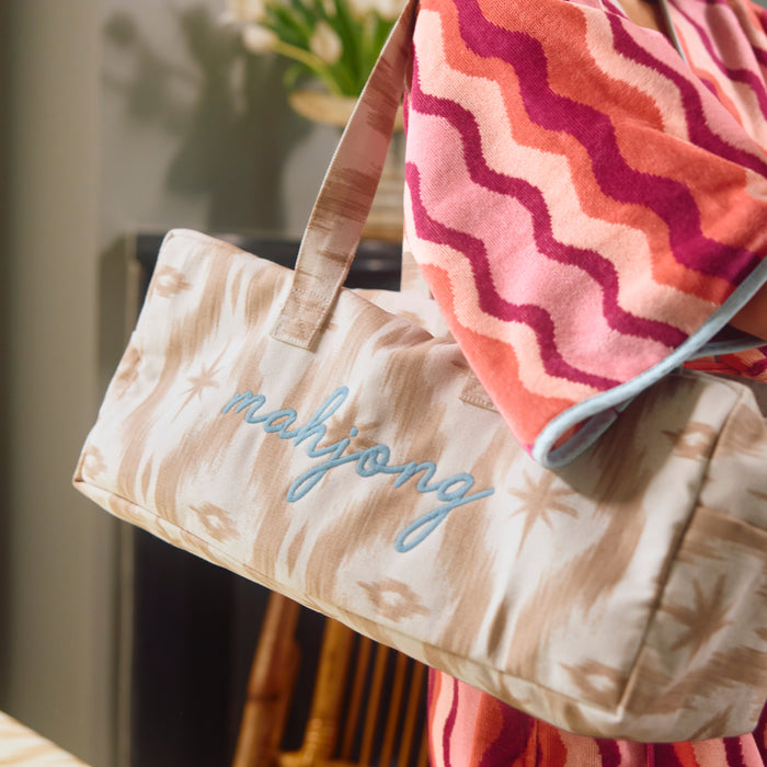 A tan and cream ikat Mahjong bag with light blue 'mahjong' embroidery, draped with a pink and red wavy-patterned towel.