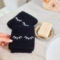 Two black towels with white embroidered closed-eye designs are shown on a light marble countertop next to a bar of soap on a small dish. A hand is holding the front towel.