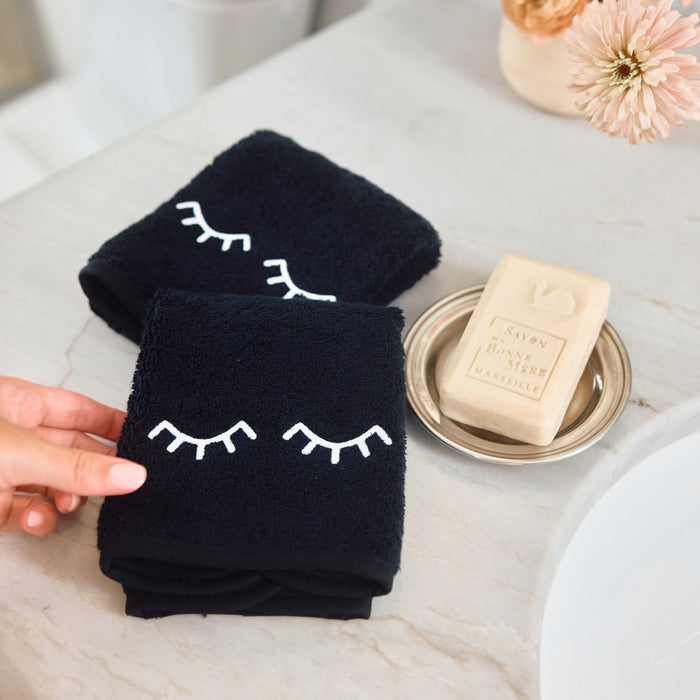 Two black towels with white embroidered closed-eye designs are shown on a light marble countertop next to a bar of soap on a small dish. A hand is holding the front towel.
