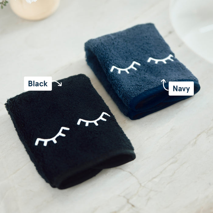 Two folded plush towels on a light marble surface, labeled Black and Navy, each with a white embroidered closed-eye design.