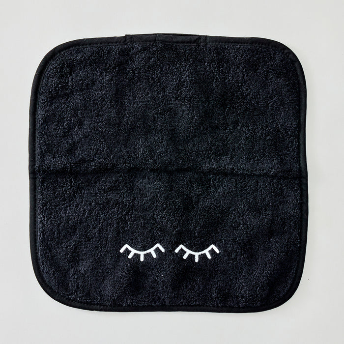 A black textured towel with rounded corners and a small white embroidered pair of closed eyelashes near the bottom center, shown flat against a light gray background.