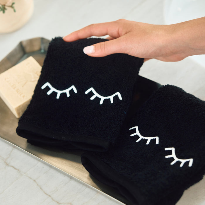 A hand holds a folded black towel with white embroidered closed-eyes motif, alongside another matching towel on a metal tray in a bright bathroom setting.