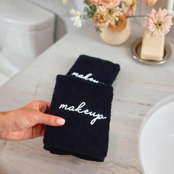 A hand holds a black terry cloth makeup towel with white cursive embroidery reading 'makeup' in a bathroom setting with a marble countertop, flowers, and a sink in the background.