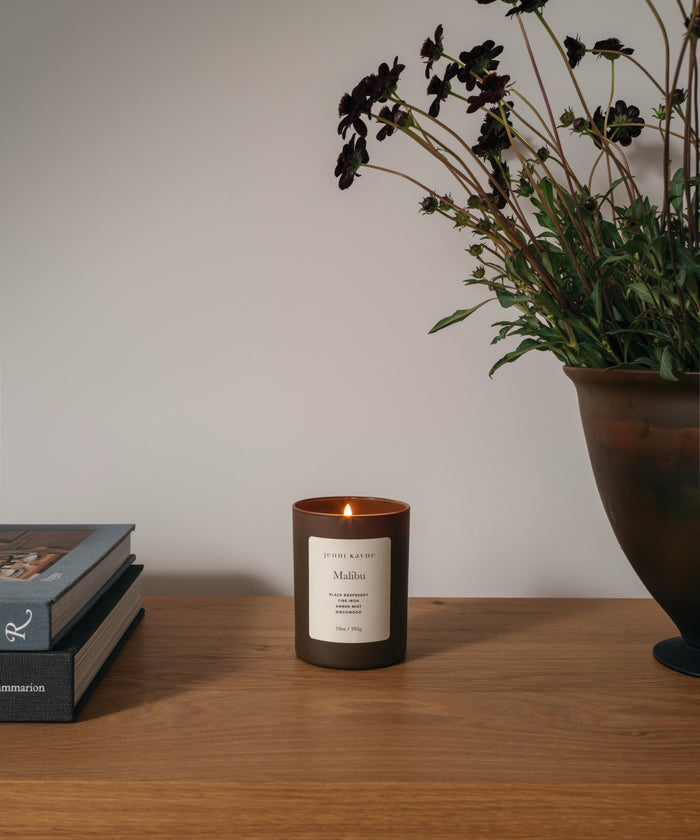 A lit candle in a dark brown glass jar sits on a wooden table, styled with stacked books on the left and a large potted plant on the right against a neutral light gray wall.
