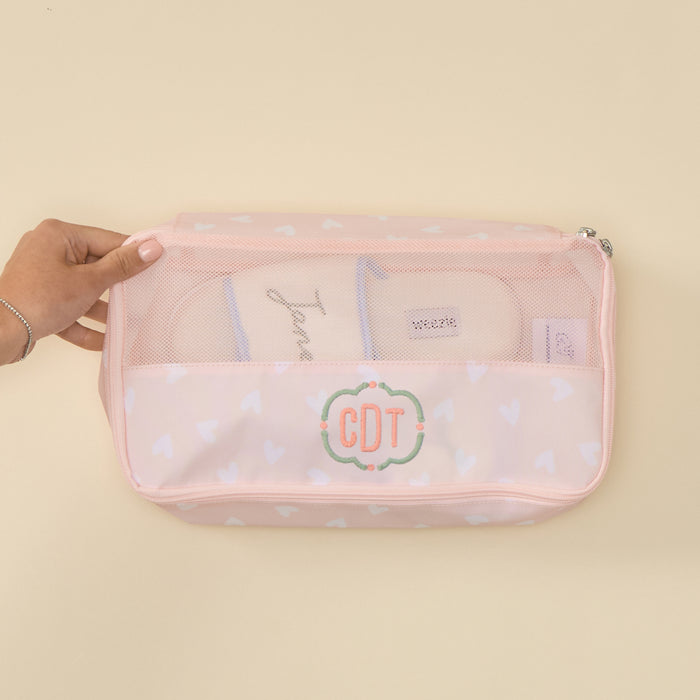 Top-down view of a rectangular, zippered Medium Packing Cube in a "Tossed Pink Hearts" print. The cube features a clear mesh top section and a pale pink lower section patterned with white hearts. The lower front panel is embroidered with the initials "CDT" in green, cursive thread inside a decorative scalloped border. A pair of beige slippers is visible inside through the mesh, and a hand is shown holding the edge of the cube against a solid, light-toned background.