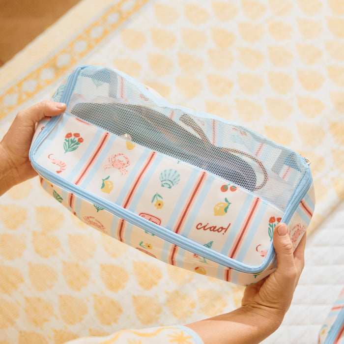 Close-up, angled view of a hands holding the "Dolce Vita" print Medium Packing Cube. The cube is patterned with small illustrations of lemons, tomatoes, flowers, and seashells on a striped background. The word "ciao!" is embroidered in red, cursive thread on the lower front panel. The top of the cube is made of blue-trimmed clear mesh, and the background shows a soft, patterned fabric surface.