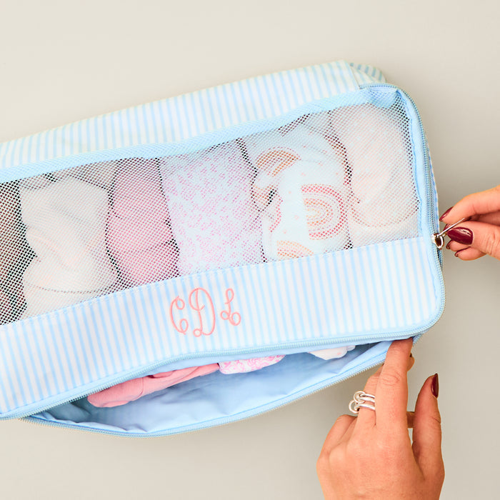 Close-up, top-down view of a rectangular, zippered Medium Packing Cube in a "Heritage Blue Stripe" print. The cube features a clear mesh top and light blue and white striped fabric sides. A hand is shown opening the zipper, revealing neatly folded clothing inside. The lower front panel is embroidered with the initials "CDL" in coral-colored, cursive thread.