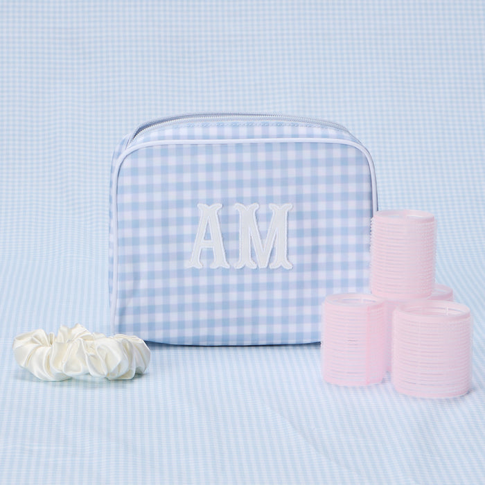 A medium, light blue gingham patterned toiletry bag with "AM" embroidered in white, sitting next to pink hair rollers.