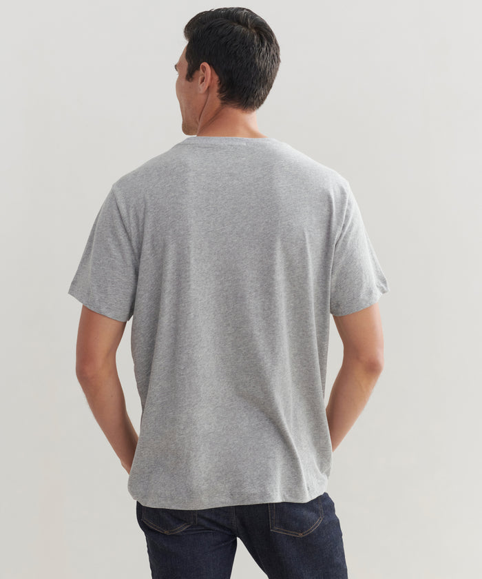 A man stands with his back to the camera, wearing a Mens Vintage Pocket Tee in grey and dark blue jeans against a light-colored background.