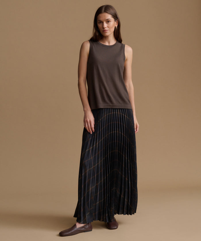 A woman modeling a sleeveless dark brown top and a long pleated skirt with thin gold stripes, standing against a solid tan studio background.