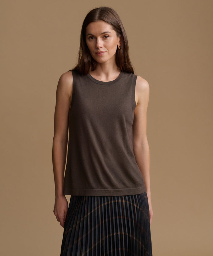 A woman modeling a sleeveless crew-neck knit top in a dark taupe-brown color, paired with a pleated dark skirt, set against a solid warm beige studio background.