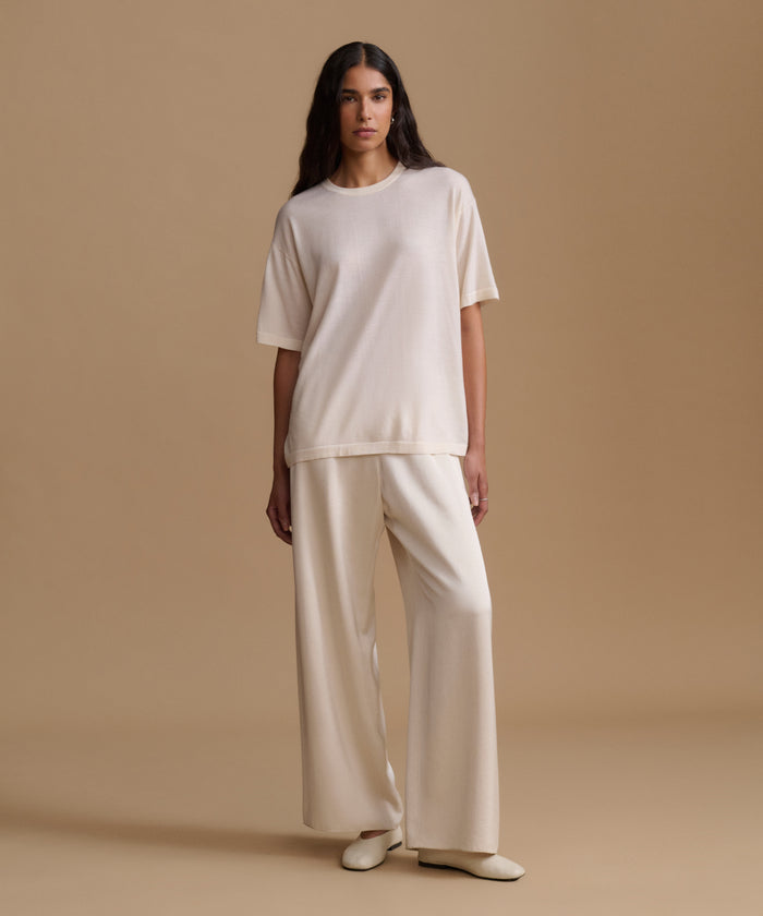 A woman modeling a matching cream-colored set consisting of a short-sleeve crewneck top and wide-leg trousers, paired with cream pointed-toe flats, set against a solid beige studio backdrop.