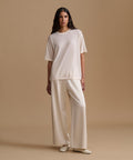 A woman modeling a matching cream-colored set consisting of a short-sleeve crewneck top and wide-leg trousers, paired with cream pointed-toe flats, set against a solid beige studio backdrop.