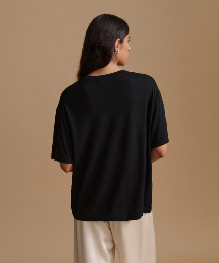 A woman viewed from the back wearing an oversized black short-sleeve t-shirt and cream-colored pants, set against a solid warm beige studio background.
