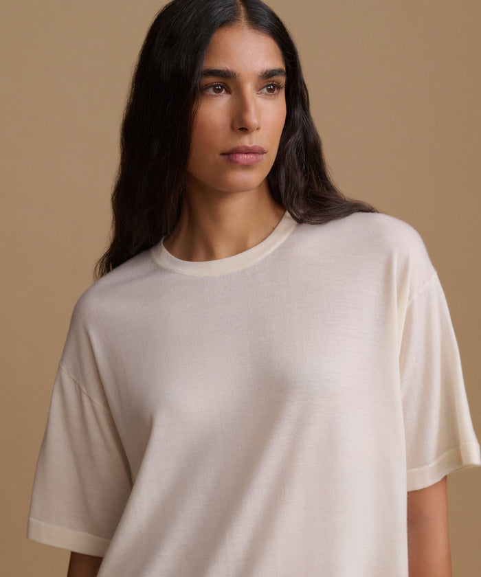 A woman wearing a cream-colored, short-sleeve crewneck t-shirt, photographed against a solid beige background.