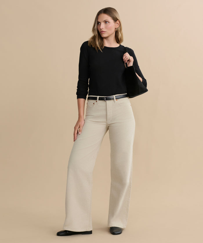 A woman stands against a beige background wearing a black long-sleeve top, the Mid Rise Wide Leg pants in light beige, a black belt, and black shoes, with one hand resting on her hip.
