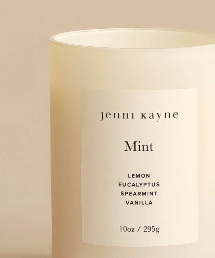 The Mint Glass Candle features a white minimalist label reading jenni kayne Mint and notes of lemon, eucalyptus, spearmint, and vanilla. Hand-poured in Los Angeles, this natural 10oz (295g) candle is set against a neutral beige background.