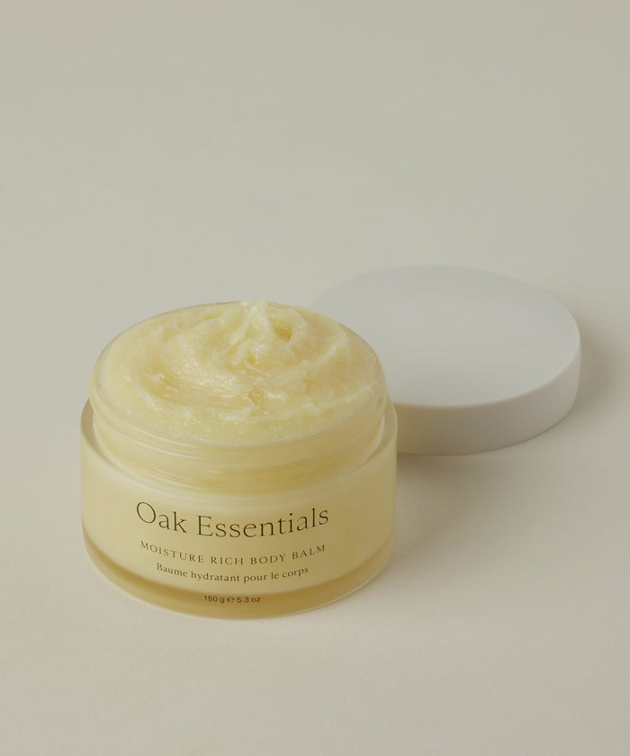 A glass jar of Moisture Rich Body Balm with a white lid sits on a light beige background, revealing its creamy, whipped texture enriched with cocoa butter inside.
