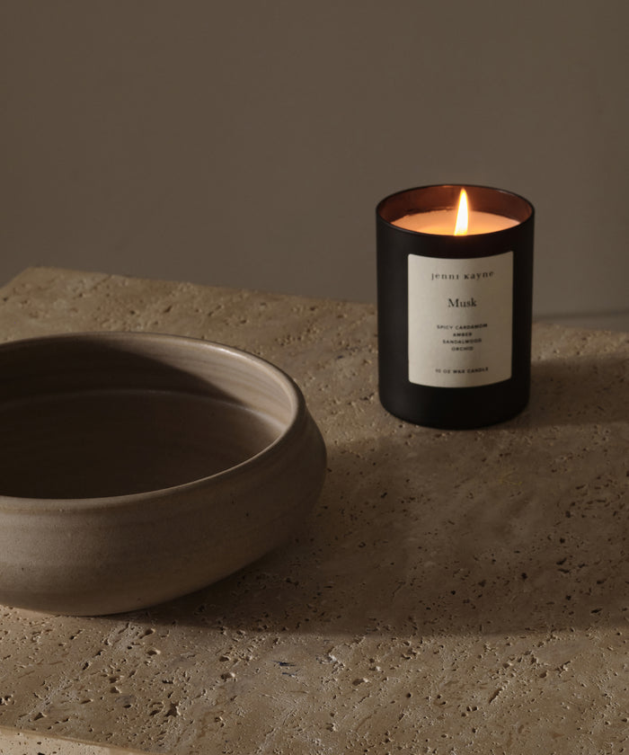 The Musk Glass Candle, hand-poured in Los Angeles with coconut soy wax, rests on a textured stone surface next to a neutral ceramic bowl, creating a calm and minimalist scene.