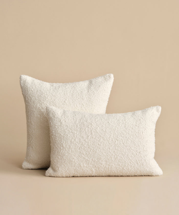 Two off-white textured throw pillows, featuring the Nell Boucle Lumbar Pillow, are arranged on a plain beige surface with a matching background; one is slightly larger and set behind the other.
