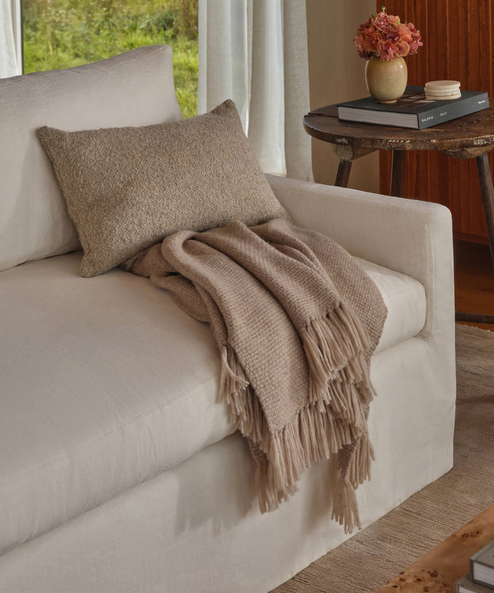 A beige sofa features a Nell Boucle Lumbar Pillow and a fringed tan throw. A round wooden side table displays stacked books and a vase of pink flowers, all illuminated by natural light from the window.