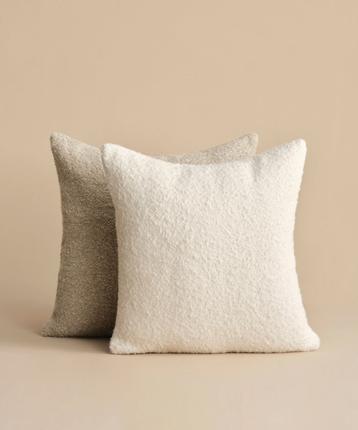The Nell Boucle Pillow, featuring a soft ivory boucle texture, is displayed in front of a light brown textured pillow against a beige background.