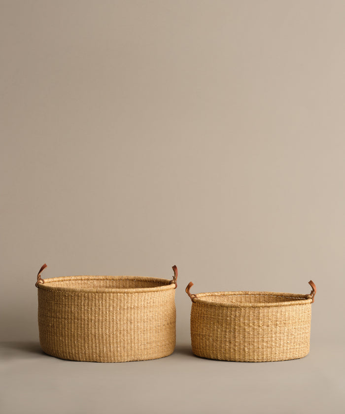 Two Nesting Baskets of different sizes, handwoven from elephant grass with tan hues and leather handles, are displayed side by side against a plain beige background.