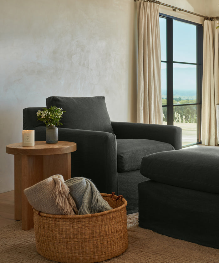 A cozy living room corner features a dark armchair, ottoman, wooden side table with a plant and candle, and a Nesting Basket filled with folded blankets near large windows dressed in beige curtains.