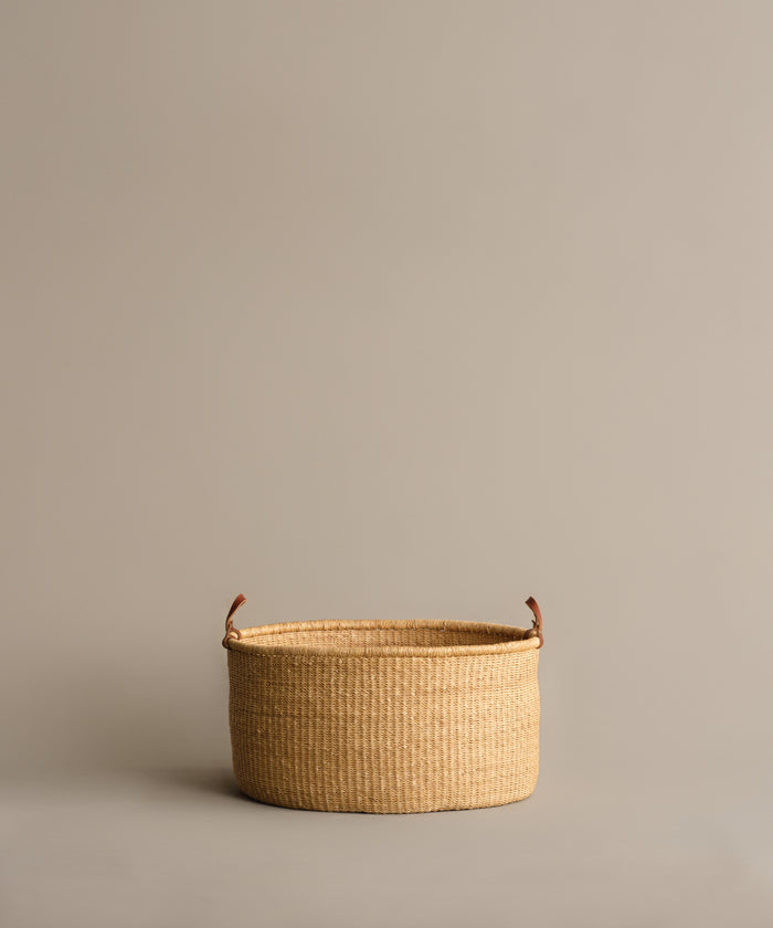 The Nesting Basket, a round handwoven elephant grass basket with brown handles, rests on a plain light beige surface against a matching beige background.