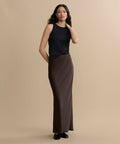 A woman stands against a beige background wearing a sleeveless black top, black shoes, and the Nina Slip Skirt in brown. She has long wavy hair, with one hand at her side and the other touching her hair.