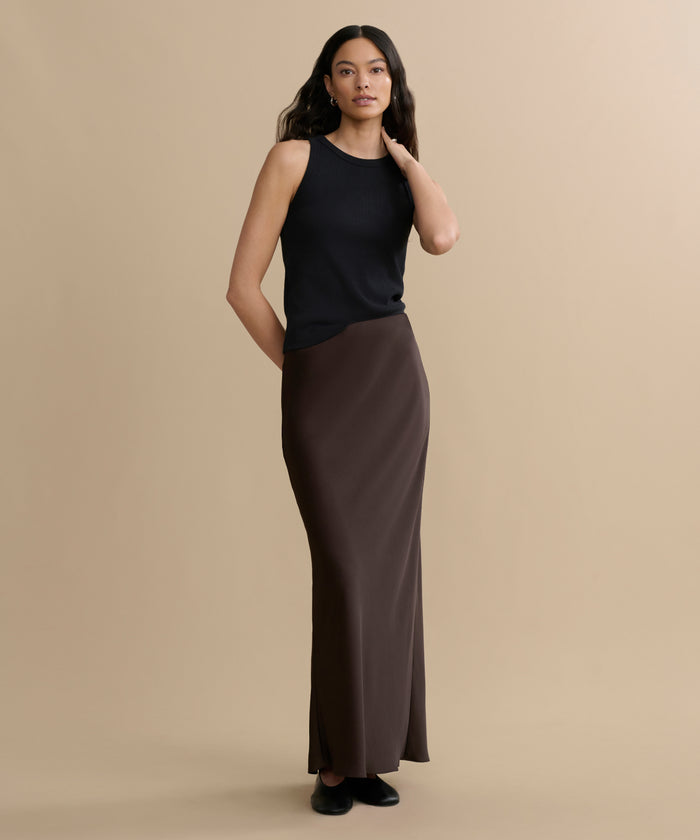 A woman stands against a beige background wearing a sleeveless black top, black shoes, and the Nina Slip Skirt in brown. She has long wavy hair, with one hand at her side and the other touching her hair.
