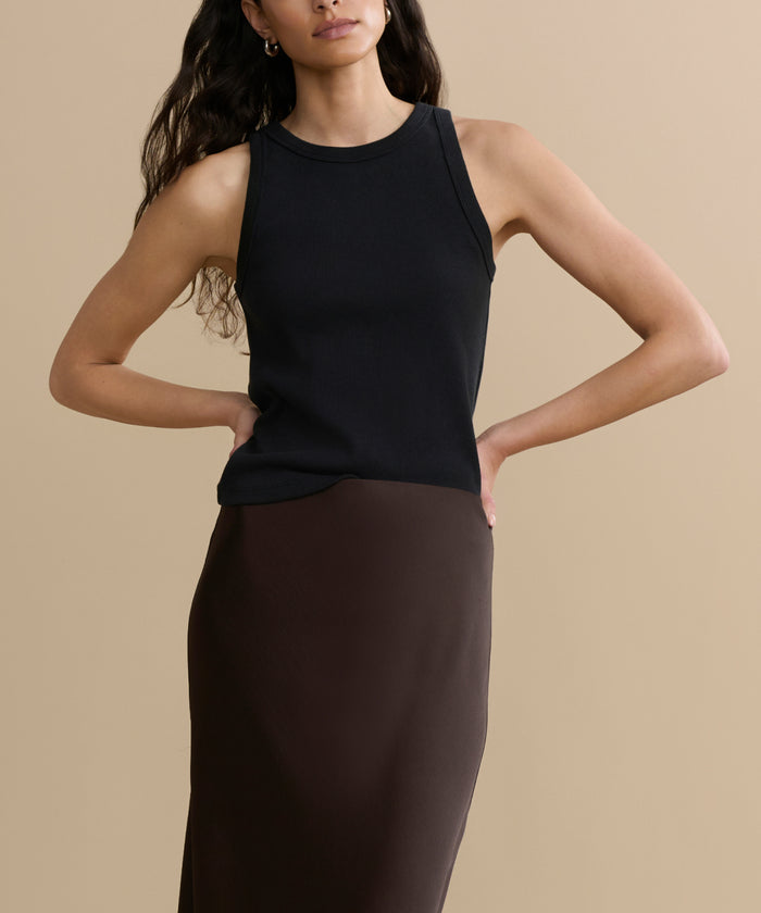 A woman with long, wavy hair wears a sleeveless black top and the high-waisted Nina Slip Skirt in dark brown. She poses against a plain beige background with her hands on her hips.