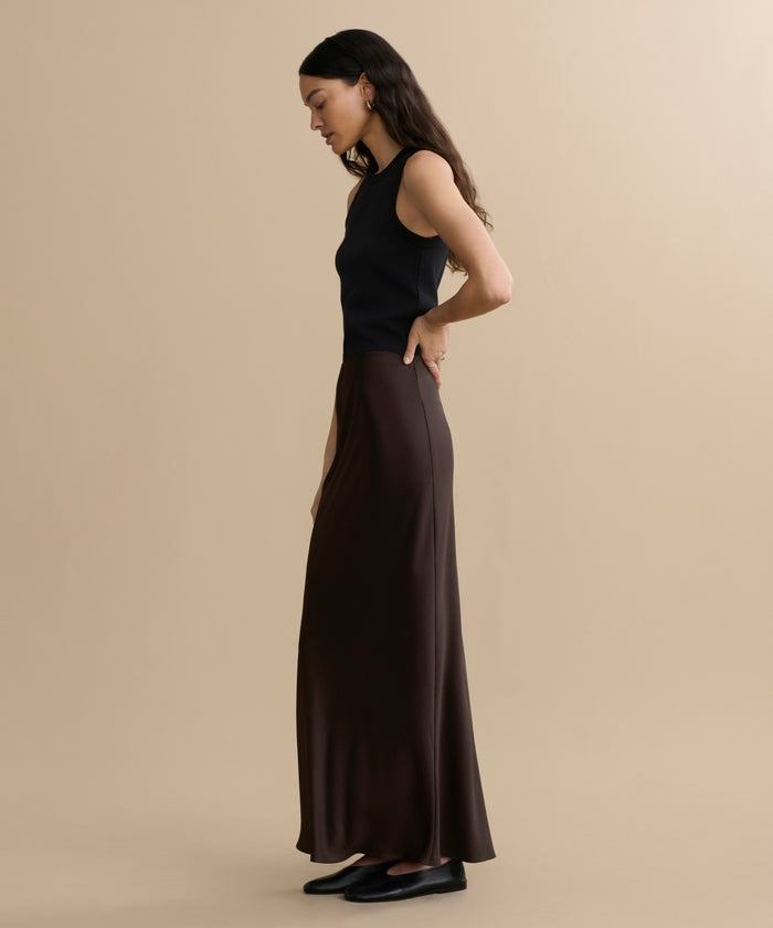 A woman with long dark hair stands in profile against a beige background, wearing a sleeveless black top, black shoes, and the Nina Slip Skirt in dark brown. One hand rests on her hip as she looks downward.