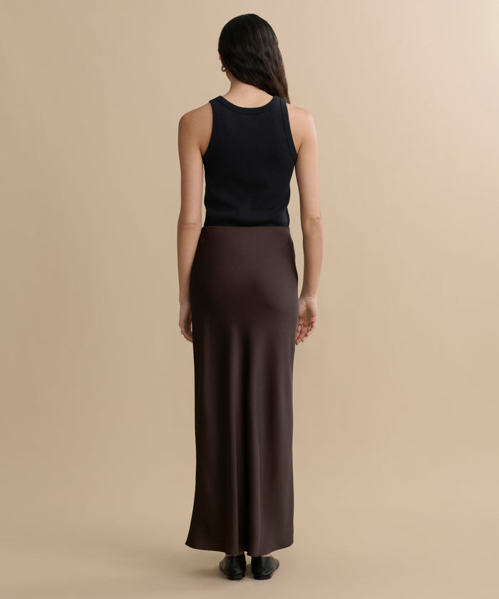 A woman with dark, wavy hair faces away from the camera against a beige background, wearing a black sleeveless top and the Nina Slip Skirt in dark brown.