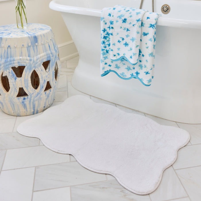 White scalloped bath rug on a tiled bathroom floor next to a tub and a blue patterned towel.