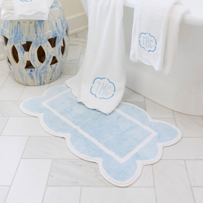 White bath rug with a light blue scalloped border next to a tub, towels, and a ceramic stool.
