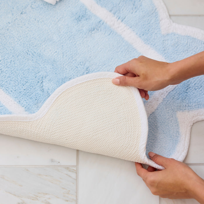 Light blue scalloped bath rug with a corner folded over to reveal beige non-slip backing.