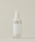 A frosted glass bottle with a white pump dispenser labeled Nourishing Cleansing Milk for sensitive skin stands against a plain, neutral background. The formula is enriched with Oat Amino Acids.