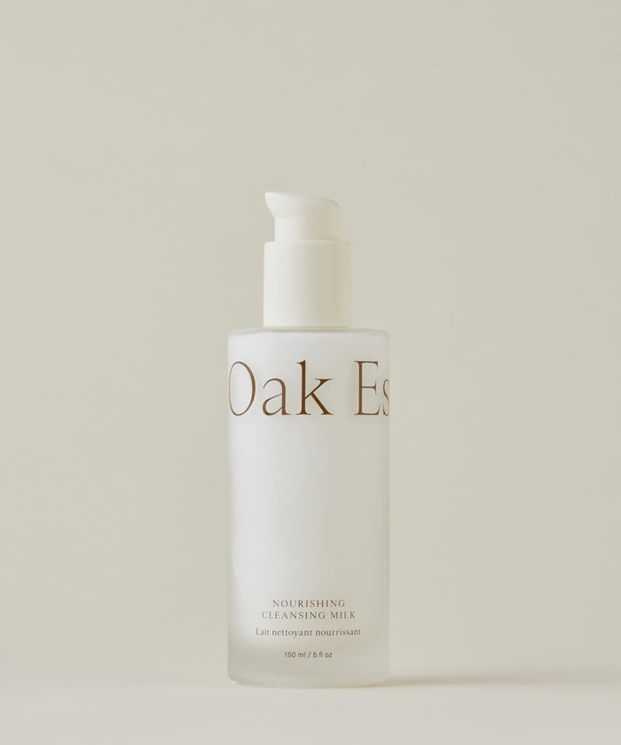 A frosted glass bottle with a white pump dispenser labeled Nourishing Cleansing Milk for sensitive skin stands against a plain, neutral background. The formula is enriched with Oat Amino Acids.